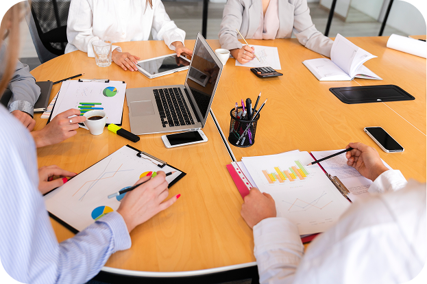 Five people sit around a conference table with laptops, charts, coffee, and documents, engaged in a business meeting or discussion.